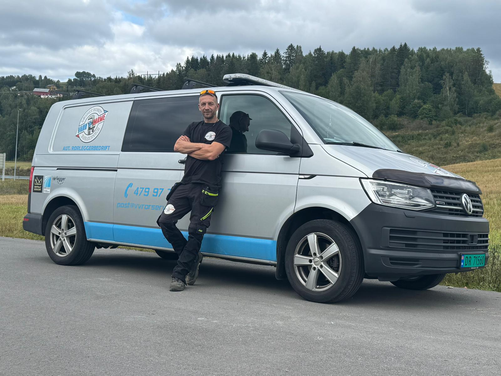 Daglig leder i Vann og Varme Rørservice AS, Darius, står avslappet inntil firmabil med logo, sertifiseringer og kontaktinformasjon på bilen